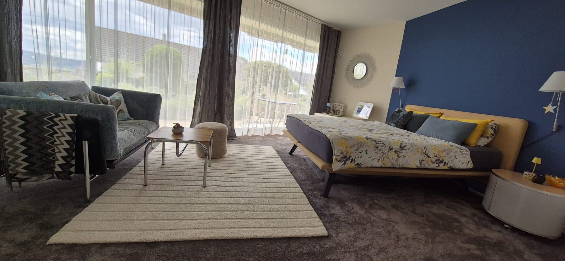 Schlafzimmer mit Bett vor blauer Wand, Sitzecke und bodentiefen Fenstern
