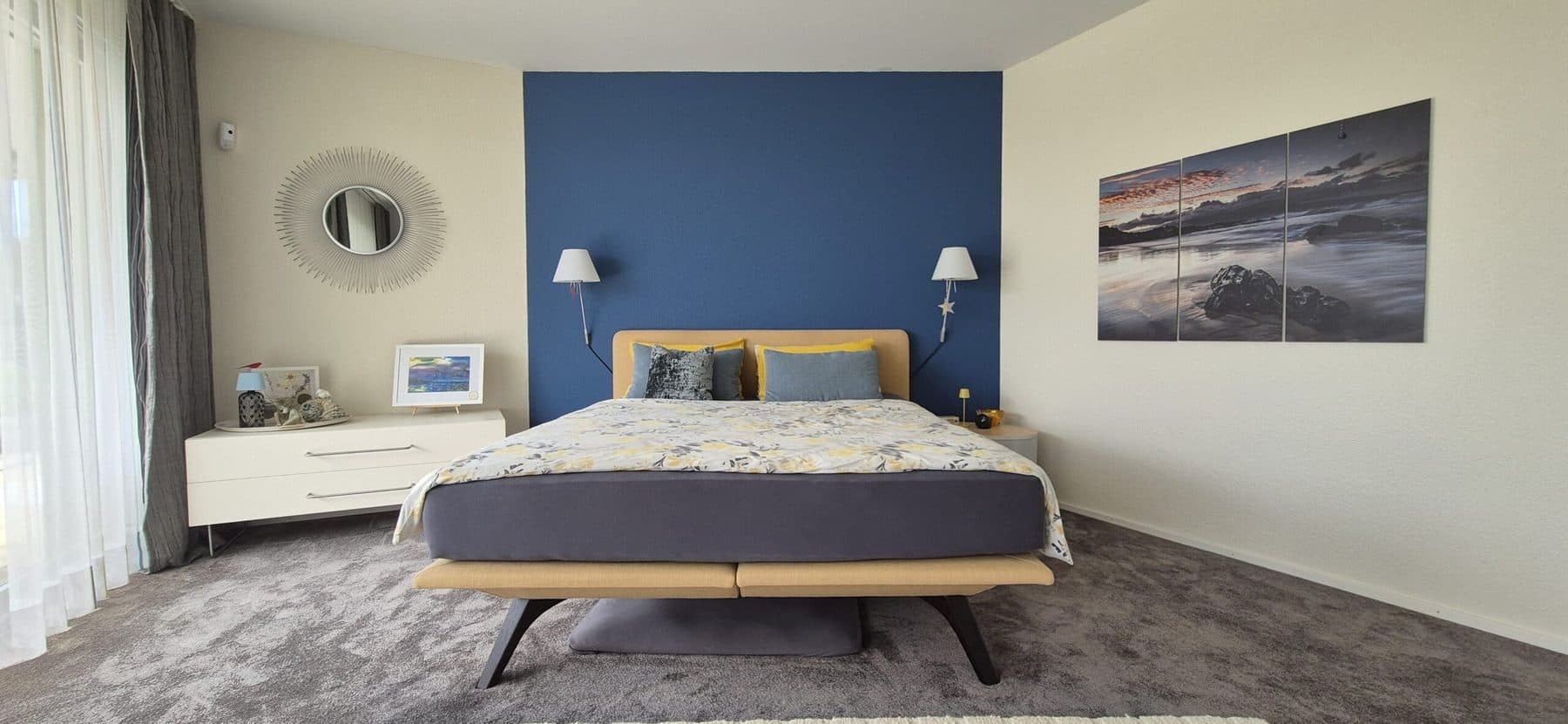 Bett vor blauem Wandakzent, Nachttischen und dreiteiligem Küstenbild